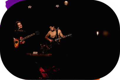 Sur une scène de concert plongée dans la pénombre deux guitaristes, une femme à gauche, un homme à droite se tiennent debout, leur instrument tenu par une bandoulière en cuir, l'air inspiré. - Agrandir l'image, fenêtre modale