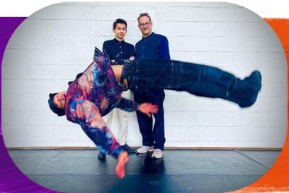 Photographie en intérieur montrant un danseur de breakdance figé en plein mouvement acrobatique horizontal (un "freeze"), suspendu en l'air au premier plan. Il porte une veste colorée et un jean large. Derrière lui, deux hommes debout, l'un plus jeune et l'un portant des lunettes, posent calmement devant un mur de briques blanches, créant un contraste saisissant avec l'action du danseur. - Agrandir l'image, fenêtre modale