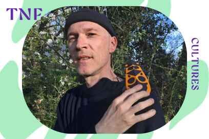 L'illustrateur jeunesse Guillaume Ruffault pos face caméra dnas un environnement naturel extérieur, pull over marin et bonnet de laine kaki sur la tête, un papillon gigantesque aux ailes fermées en papier coloré orange et noir, posé sur son épaule gauche - Agrandir l'image, fenêtre modale