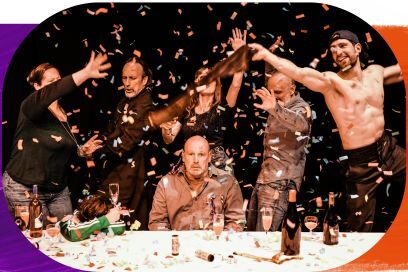 Scène théâtrale montrant un groupe de personnes en pleine fête autour d'une table recouverte de confettis multicolores. Au centre, un homme chauve regarde fixement l'objectif avec un air sérieux et hébété. Autour de lui, les autres personnages dansent, lèvent les bras et célèbrent de manière exubérante, notamment un homme torse nu sur la droite. Une pluie de confettis s'abat sur l'ensemble de la scène sur fond noir. - Agrandir l'image, fenêtre modale