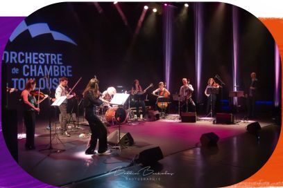 Une vue d'ensemble d'un concert sur scène de l'Orchestre de Chambre de Toulouse, dont le logo est projeté en arrière-plan à gauche. Une dizaine de musiciens — violonistes, violoncelliste, batteur, guitariste et contrebassiste — sont répartis sur la scène sous des projecteurs aux teintes roses et blanches. Au premier plan, une violoniste en combinaison noire étincelante joue de dos face au reste du groupe. - Agrandir l'image, fenêtre modale