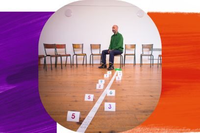 Vue toute en perspective d'un homme chauve assis sur un tabouret au milieu d'une pièce au parquet en bois. Il porte une chemise verte et un jean. À ses pieds, une longue bande de papier blanc traverse la pièce en diagonale, portant l'inscription "QUE J'AIME À FAIRE APPRENDRE CE NOMBRE UTILE AUX SAGES", entourée de petits papiers numérotés pliés en arche. L'homme lève les yeux vers l'objectif. - Agrandir l'image, fenêtre modale