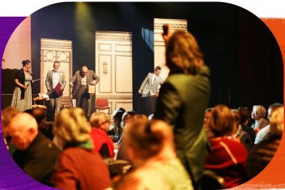 Photographie prise depuis le public lors d'une représentation théâtrale sur scène du Loto du Grenier de Toulouse.. Au premier plan, de dos et légèrement flous, les spectateurs sont assis à des tables. Sur la scène éclairée en arrière-plan, quatre comédiens en costumes évoluent devant des décors représentant des portes blanches au style classique. - Agrandir l'image, fenêtre modale
