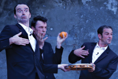 photo d'un spectacle, on voit trois hommes habillés en costume (veste noire et chemise blanche), un tient un plateau d'argent, un autre une pomme, et un autre a la main sur la poitrine et l'air perplexe exagéré