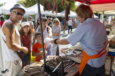 photo d'un stand de charcuterie avec des saucissons pendus et posés sur le stand, un homme derrière le stand d'où la photo est prise tend un sachet à une femme accompagnée de deux enfants et un homme qui sourient à l'objectif - Agrandir l'image 13 sur 15, fenêtre modale