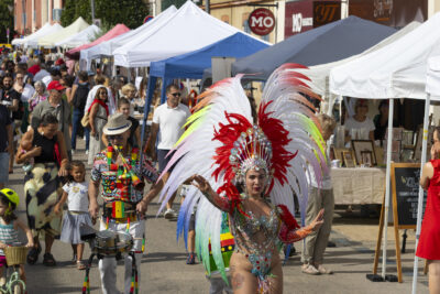photo d'un défilé avec en tête une femme habillée façon carnaval, dans une rue commerçante ensoleillée - Agrandir l'image 12 sur 15, fenêtre modale