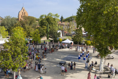 photo de la place de la mairie avec beaucoup d'arbres et de verdure, la façade de l'église à l'arrière-plan à gauche, et un spectacle de danse tahitienne sur une scène sur le parvis - Agrandir l'image 7 sur 15, fenêtre modale