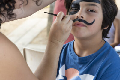 photo d'un jeune garçon qui se fait maquiller une moustache noire, et porte déjà en maquillage un cache oeil noir, façon pirate - Agrandir l'image 11 sur 15, fenêtre modale