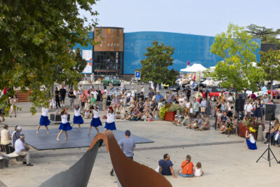 photo du parvis de la mairie avec vue sur la scène où a lieu un spectacle de danse tahitienne, une cinquantaine de personnes rassemblées devant, à l'arrière-plan, on voit la façade de l'Escale, bâtiment grand et bleu clair - Agrandir l'image 6 sur 15, fenêtre modale