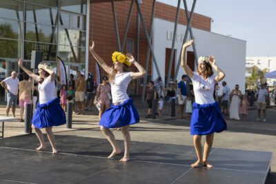 photo d'une animation de danse tahitienne sur la scène en extérieur, trois femmes avec jupe bleue et tee-shirt blanc dansent, quelques personnes avec enfants les regardent depuis l'ombre en arrière-plan - Agrandir l'image 10 sur 16, fenêtre modale