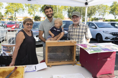 photo de trois personnes avec un bébé blond, au centre un homme tient le bébé dans les bras, ils sont derrière un stand sur lequel il y a une ruche sous verre avec des abeilles visibles - Agrandir l'image 9 sur 16, fenêtre modale