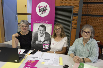 photo du stand des Restaurants du coeur, trois femmes assises derrière, deux tiennent le portrait noir et blanc de Coluche - Agrandir l'image 15 sur 16, fenêtre modale