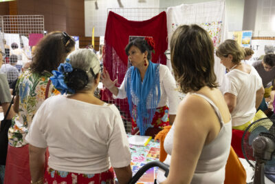 photo d'un stand avec une femme derrière qui porte un châle bleu, façon andalouse, et trois femmes devant qui l'écoutent parler - Agrandir l'image 14 sur 16, fenêtre modale