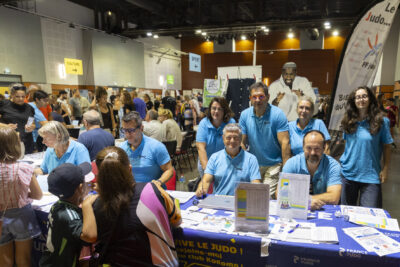 photo du stand de judo, en intérieur dans la grande salle, une dizaine de personnes derrière, habillées en bleu clair, et devant le stand une femme et un enfant qui regardent - Agrandir l'image 12 sur 16, fenêtre modale