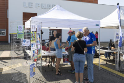 photo d'un stand extérieur avec des adultes qui échangent, habillés en bleus, sous un barnum. Derrière, on voit un grand bâtiment avec façade blanche et ocre - Agrandir l'image 5 sur 16, fenêtre modale