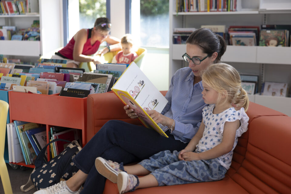 photo d'une femme qui lit une histoire à une jeune fille dans un canapé, et à l'arrière plan, une femme qui montre un livre à un enfant dans un fauteuil