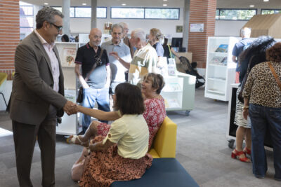 photo du Maire et d'une dizaine de personnes dans la Médiathèque, lumineuse, le Maire serre la main à une femme assise sur un fauteuil - Agrandir l'image 13 sur 19, fenêtre modale