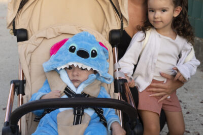 photo d'une famille, une femme à genoux à côté d'une petite fille, et d'une poussette dans laquelle il y a un bébé déguisé en Stitch - Agrandir l'image 10 sur 25, fenêtre modale
