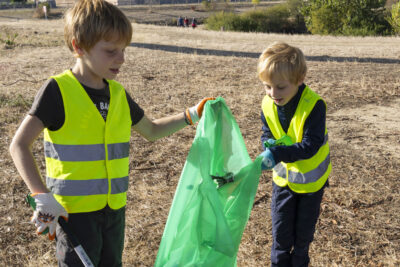 photo de deux enfants, un tien un sac poubelle vert, un autre y jette un déchet, ils sont dehors dans une sorte de parc, l'herbe est sèche - Agrandir l'image 9 sur 9, fenêtre modale