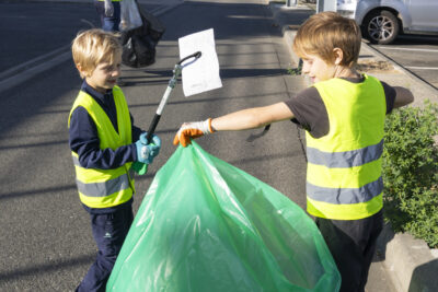photo de deux enfants en gilet jaune, un porte un grand sac poubelle vert, l'autre s'apprête à y jeter un bout de carton, ils sont sur un parking avec un adulte qui les regarde plus loin, lui aussi en gilet jaune - Agrandir l'image 7 sur 9, fenêtre modale