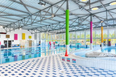 photo de l'intérieur d'une piscine, avec vue sur un grand bassin avec 5 couloirs de nage, un sol bleu et blanc, des poteaux colorés pour soutenir le toit. L'espace est grand et lumineux