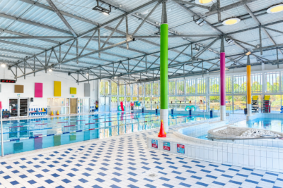 photo de l'intérieur d'une piscine, avec vue sur un grand bassin avec 5 couloirs de nage, un sol bleu et blanc, des poteaux colorés pour soutenir le toit. L'espace est grand et lumineux - Agrandir l'image 2 sur 3, fenêtre modale