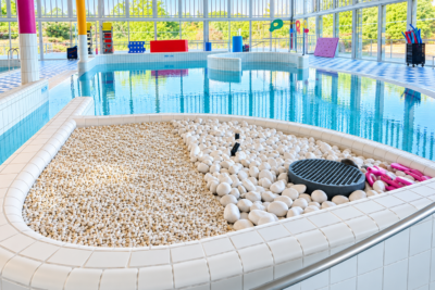 photo d'un petit bassin de piscine, avec au premier plan une sorte de bac avec galets décoratifs blancs, et un bassin très propre avec personne, autour de grandes baies vitrées qui donnent sur la nature - Agrandir l'image 1 sur 3, fenêtre modale