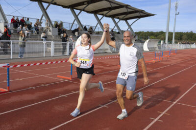 photo paysage d'un homme et d'une jeune femme qui courent sur une piste d'athlétisme, ils se tiennent la main en l'air et sourient. Derrière eux, on voit les gradins avec une vingtaine de personnes rassemblées - Agrandir l'image 12 sur 19, fenêtre modale