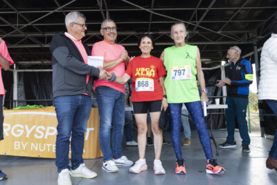 photo de deux femmes plutôt âgées en tenues de sport et avec le dossard collé sur le tee-shirt; une serre la main à un homme sur la gauche de la photo qui sourit et tient une enveloppe, un autre homme à côté sourit et applaudit, habillé d'un tee-shirt rose - Agrandir l'image 14 sur 19, fenêtre modale