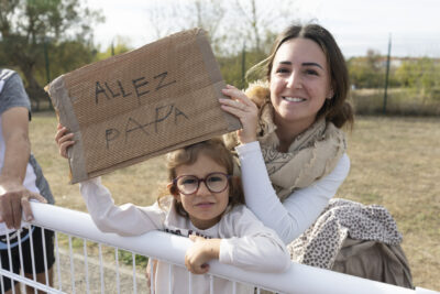 Photo paysage d'une femme et d'une petite fille qui tiennent une pancarte en carton avec écrit "Allez papa", elles sont derrière une barrière blanche et sourient à l'obejctif - Agrandir l'image 16 sur 19, fenêtre modale