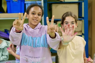 photo de deux jeunes filles qui posent pour l'objectif, une fait signe peace avec les mains, l'autre tend la main avec traces de peintures - Agrandir l'image 3 sur 18, fenêtre modale