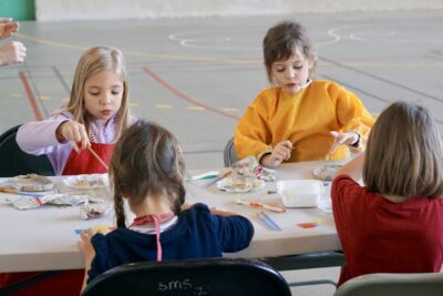 photo de quatre enfants à une table rectangulaire, deux de dos, deux de face, en train de décorer des objets de poterie gris - Agrandir l'image 17 sur 18, fenêtre modale