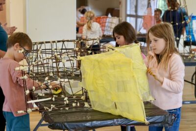 photo de trois enfants autour d'une grande lanterne en bois, occupés à mettre du papier jaune clair et transparent autour, dans une salle avec d'autres enfants qu'on distingue de loin à l'arrière plan en train de faire la même chose - Agrandir l'image 1 sur 18, fenêtre modale