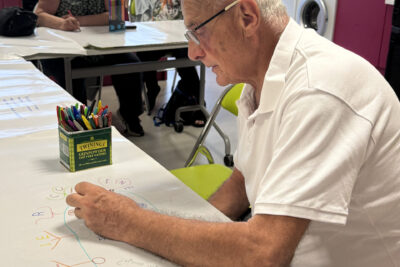 Photo d'un homme âgé assis en train de dessiner à une table, derrière lui deux femmes font la même chose - Agrandir l'image 3 sur 5, fenêtre modale