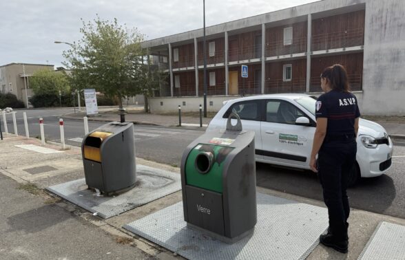 Photo dans la rue, d'une femme de biais avec un uniforme bleu foncé sur lequel il est écrit A.S.V.P., à côté de deux bacs de dépôt et d'une voiture blanche arrêtée sur la rue