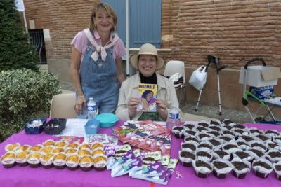 photo d'un stand couvert de muffins, avec derrière deux femmes, une assise une debout, assise, elle tient un document visuel avec écrit "ENSEMBLE" et trois illustrations de femmes - Agrandir l'image 8 sur 16, fenêtre modale