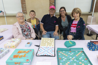 photo d'un stand de jeu avec notamment un plateau de scrabble ouvert, derrière cinq personnes dont un enfant assises qui sourient à l'objectif - Agrandir l'image 10 sur 16, fenêtre modale