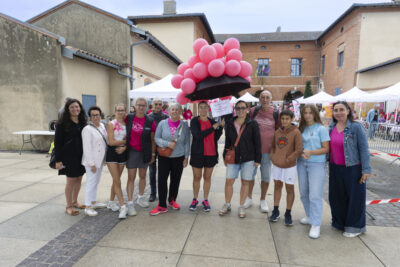 photo d'un groupe de personnes rassemblées sur le parvis de la mairie, en ligne, pour la plupart habillées en rose, sous un temps gris, toutes sourient à l'objectif et au centre une femme tient un parapluie sur lequel sont accrochés de nombreux ballons roses - Agrandir l'image 12 sur 16, fenêtre modale