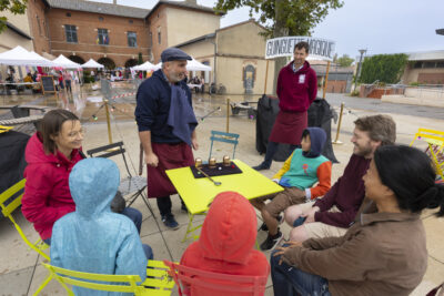 photo d'un espace jeu sur le parvis de la mairie, avec une table verte autour de laquelle 6 personnes sont réunies, dont des enfants, et un homme fait face, derrière il y a une pancarte "Guinguette magique", le temps est pluvieux - Agrandir l'image 13 sur 16, fenêtre modale