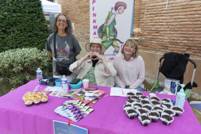 photo d'un stand en extérieur, avec une table couverte d'une nappe rose, avec des muffins et des documents, derrière le stand, trois femmes qui sourient à l'objectif - Agrandir l'image 16 sur 16, fenêtre modale