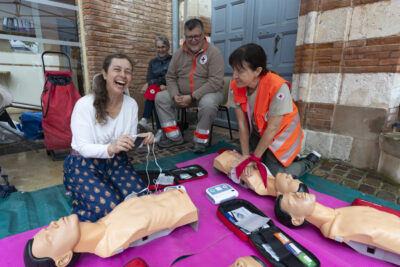 photo de quatre personnes, deux femmes agenouillées en train de pratiquer un massage cardiaque et d'utiliser un défibrillateur sur des mannequins, derrière un homme et une femme assis, ils sont en extérieur - Agrandir l'image 3 sur 16, fenêtre modale