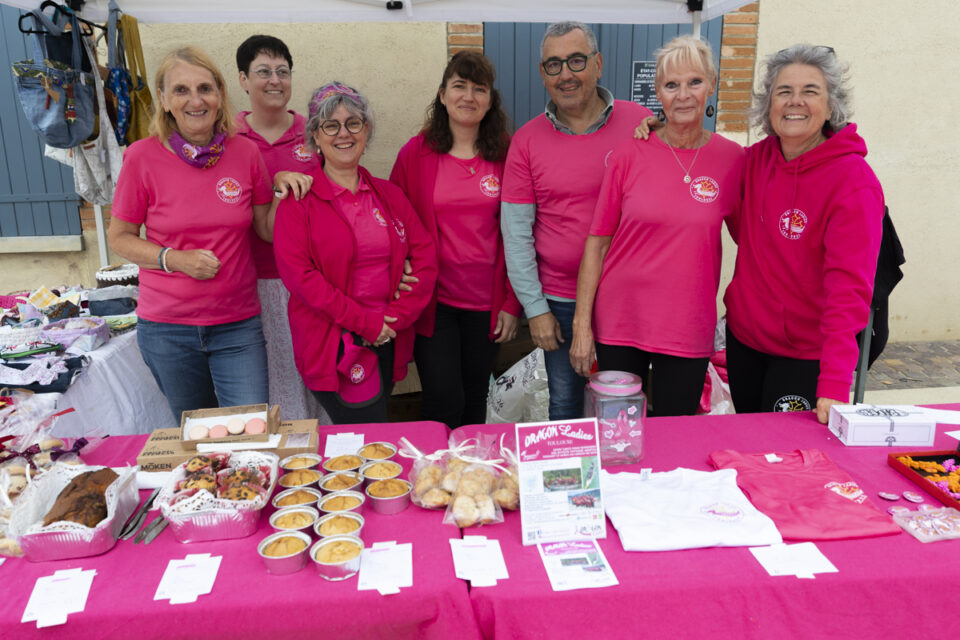 photo de sep personnes habillées d'un tee-shirt rose derrière un stand rose sur lequel il y a des pâtisseries et gâteaux en vente
