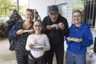 photo d'une famille avec une femme et un homme, et deux jeunes filles qui sourient à l'objectif en mangeant quelque chose dans une assiette en carton, en extérieur - Agrandir l'image 2 sur 11, fenêtre modale