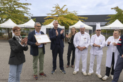 photo du Maire qui tient et montrer une sorte de diplôme sur lequel on voit un dessin de trois carottes, à côté les gens applaudissent, sur la droite des cuisiniers en tenue de travail, tous sont en extérieur - Agrandir l'image 6 sur 11, fenêtre modale