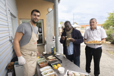 photo d'un stand avec un homme en tablier derrière qui sourit à l'objectif, et devant un homme qui goûte quelque chose dans une mini assiette, et un autre qui porte à manger sur un plateau - Agrandir l'image 4 sur 11, fenêtre modale