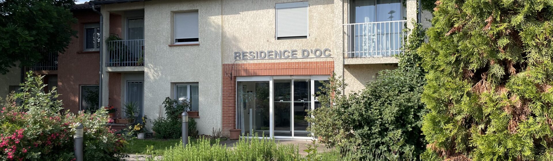 photo de la façade de la résidence d'Oc vue depuis un bosquet fleuri à côté de l'allée qui mène à l'entrée, le ciel est ensoleillé