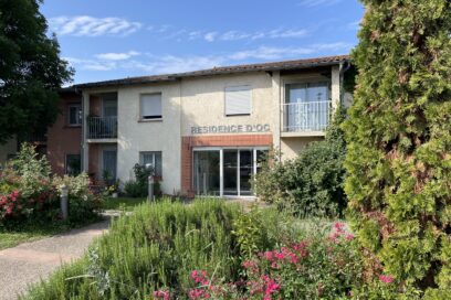 photo de la façade de la résidence d'Oc vue depuis un bosquet fleuri à côté de l'allée qui mène à l'entrée, le ciel est ensoleillé - Agrandir l'image, fenêtre modale