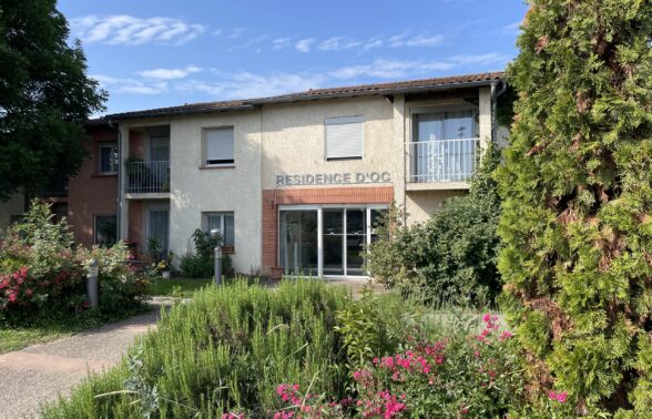 photo de la façade de la résidence d'Oc vue depuis un bosquet fleuri à côté de l'allée qui mène à l'entrée, le ciel est ensoleillé