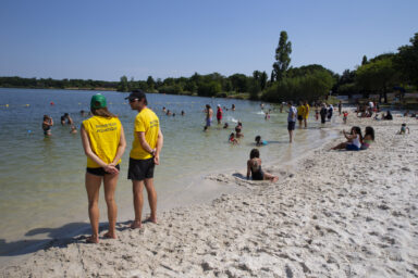Photo de la plage du lac de la Ramée, 2 sauveteurs aquatiques au premier plan, des gens qui se baignent et allongés sur la plage derrière