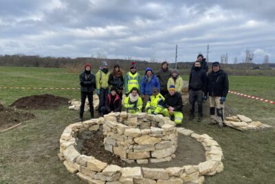 Groupe de personnes avec gilets jaunes réunis derrière une structure naturelle terre entourée de pierres - Agrandir l'image 7 sur 8, fenêtre modale